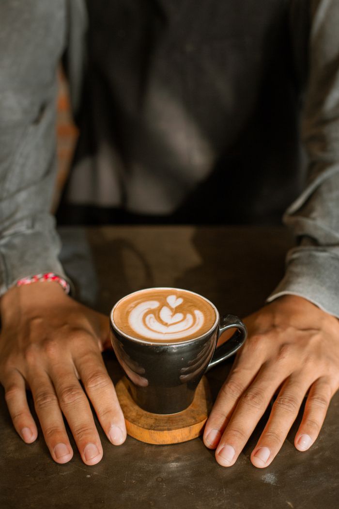 get-in-touch Warm coffee with intricate latte art held on a table, framed by relaxed hands.