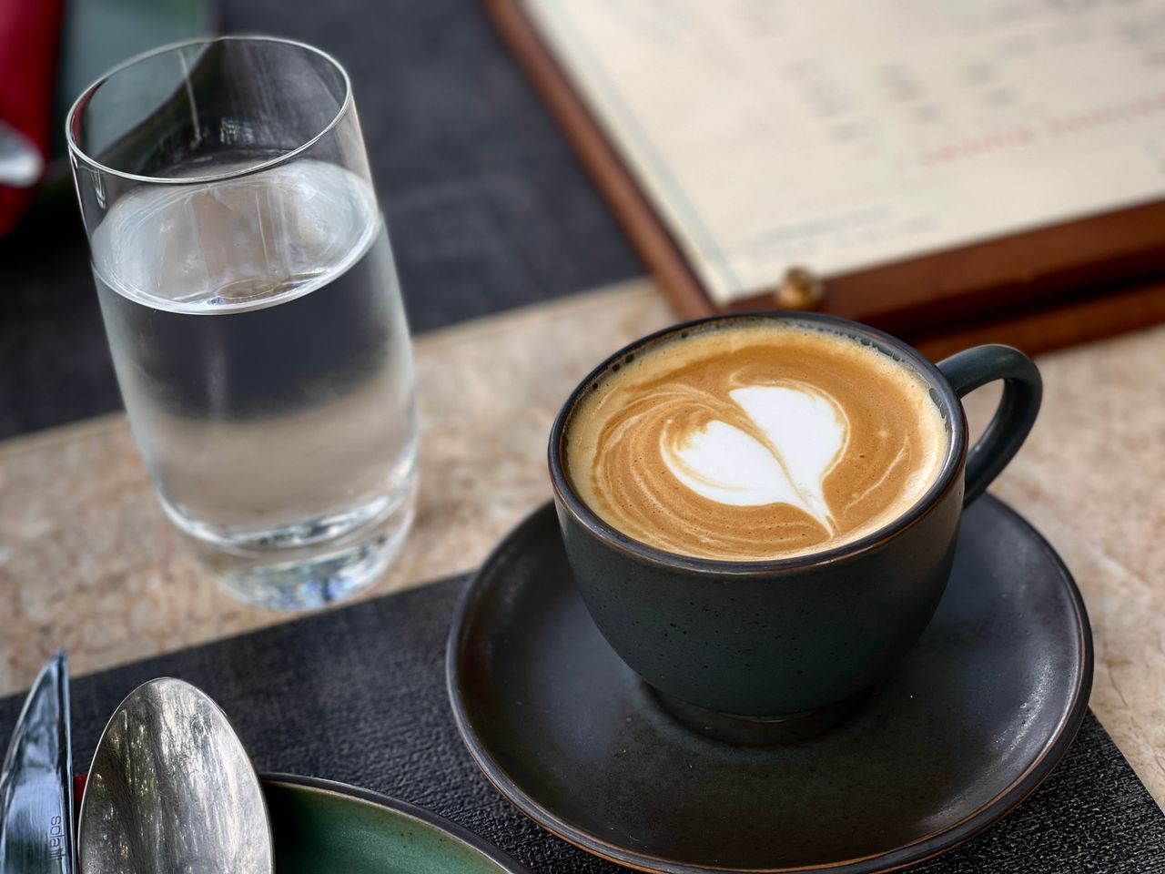 services-bg Close-up of a latte with heart-shaped latte art beside a glass of water on a cafe table.